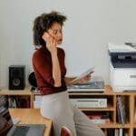 Xerox® C325 Farb-Multifunktionsdrucker in einer Home-Office-Umgebun Eine Frau sitzt an einem Schreibtisch und telefoniert neben einem Xerox® C325-Drucker