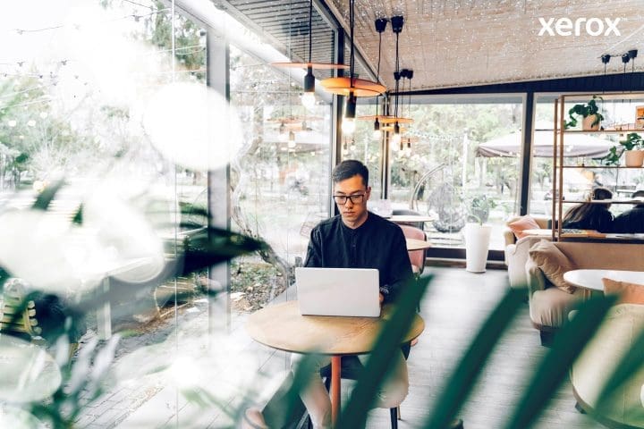 Ein Mann arbeitet fleißig an seinem Laptop in einem geschäftigen Café und ist inmitten einer lebhaften Umgebung in seine Aufgaben vertieft.