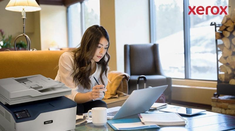 Eine Frau arbeitet in einem gemütlichen Büro oder einer Lounge an einem Laptop. Im Vordergrund ist ein kompakter Xerox-Drucker zu sehen, der auf eine nahtlose Integration des Druckens in digitale Arbeitsabläufe hinweist.